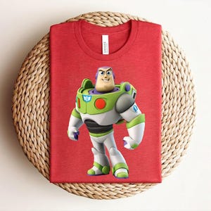 May include: Red t-shirt featuring a graphic of Buzz Lightyear from Toy Story. The character is depicted in his signature green, white, and purple spacesuit. The shirt is folded on a woven, circular surface.