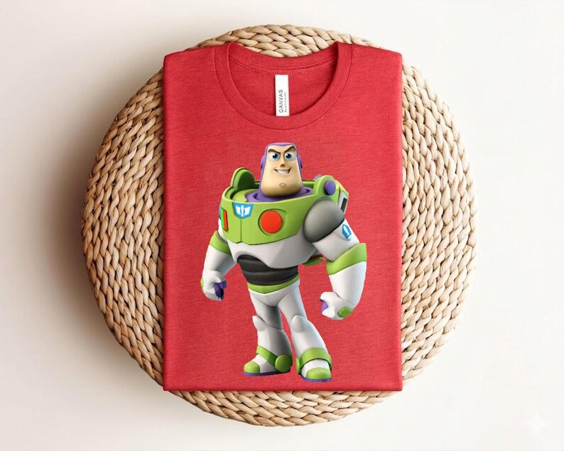May include: Red t-shirt featuring a graphic of Buzz Lightyear from Toy Story. The character is depicted in his signature green, white, and purple spacesuit. The shirt is folded on a woven, circular surface.