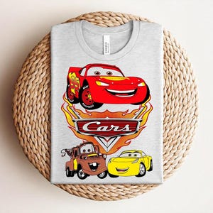 May include: A light gray t-shirt featuring a colorful graphic of characters from the animated movie "Cars." The design includes a red race car, a brown tow truck, and a yellow sports car, with the word "Cars" prominently displayed.