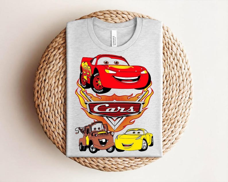 May include: A light gray t-shirt featuring a colorful graphic of characters from the animated movie "Cars." The design includes a red race car, a brown tow truck, and a yellow sports car, with the word "Cars" prominently displayed.