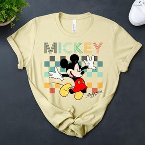 May include: A light yellow t-shirt featuring a colorful graphic of Mickey Mouse. The design includes the word "MICKEY" in a multi-colored font above a checkered pattern. Mickey Mouse is depicted with his signature red shorts, white gloves, and yellow shoes.