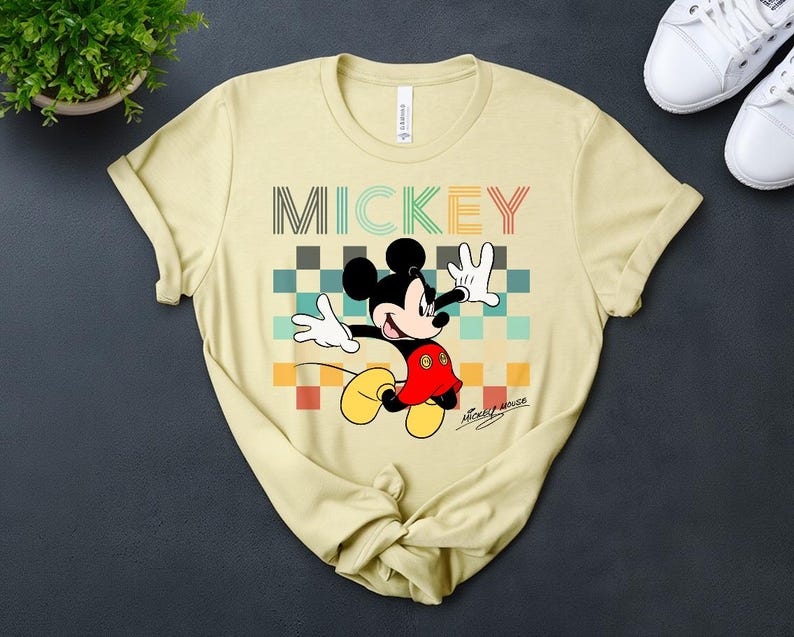 May include: A light yellow t-shirt featuring a colorful graphic of Mickey Mouse. The design includes the word "MICKEY" in a multi-colored font above a checkered pattern. Mickey Mouse is depicted with his signature red shorts, white gloves, and yellow shoes.