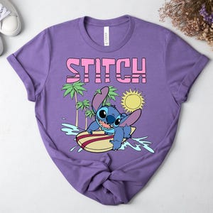 May include: A purple t-shirt featuring the word "STITCH" in pink block letters above a cartoon image of Stitch surfing. The design includes palm trees, a yellow sun, and blue water. The shirt is made of a soft material.