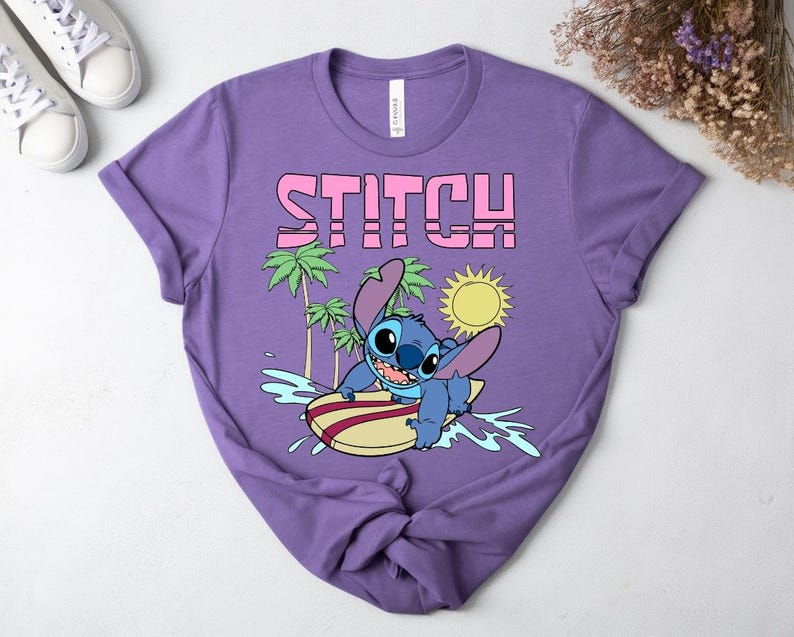 May include: A purple t-shirt featuring the word "STITCH" in pink block letters above a cartoon image of Stitch surfing. The design includes palm trees, a yellow sun, and blue water. The shirt is made of a soft material.