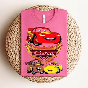 May include: Pink t-shirt featuring a graphic of Lightning McQueen, Mater, and a yellow car from the movie Cars. The Cars logo is below the cars, with flames around the logo. The shirt is folded on a woven, circular surface.