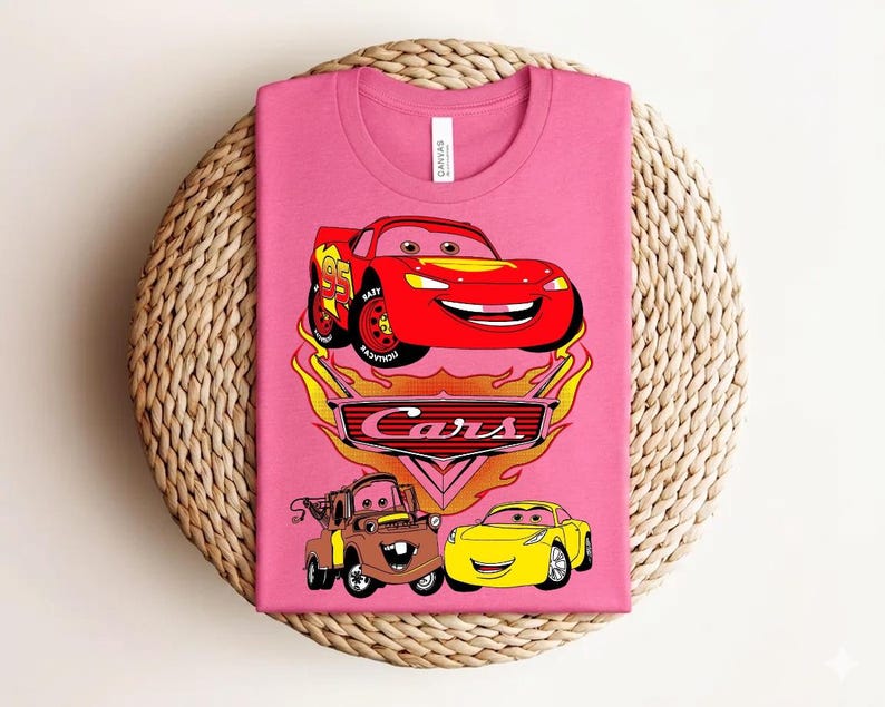 May include: Pink t-shirt featuring a graphic of Lightning McQueen, Mater, and a yellow car from the movie Cars. The Cars logo is below the cars, with flames around the logo. The shirt is folded on a woven, circular surface.