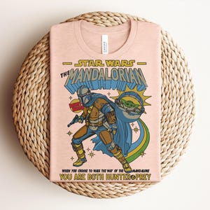 K&ouml;nnte beinhalten: Hellrosa T-Shirt mit einer Grafik von Star Wars The Mandalorian. Das Design zeigt die Mandalorianer-Figur, Baby Yoda und den Text "STAR WARS THE MANDALORIAN" und "WHEN YOU CHOOSE TO WALK THE WAY OF THE MANDALORE YOU ARE BOTH HUNTER PREY."