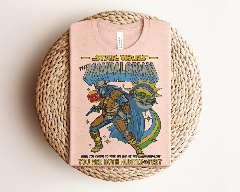 K&ouml;nnte beinhalten: Hellrosa T-Shirt mit einer Grafik von Star Wars The Mandalorian. Das Design zeigt die Mandalorianer-Figur, Baby Yoda und den Text "STAR WARS THE MANDALORIAN" und "WHEN YOU CHOOSE TO WALK THE WAY OF THE MANDALORE YOU ARE BOTH HUNTER PREY."