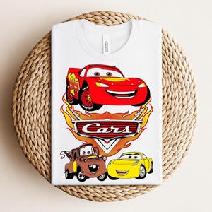 May include: White t-shirt featuring a graphic of the Cars movie characters Lightning McQueen, Mater, and a yellow car. The design includes the word "Cars" in a flame-like font. The shirt is folded on a woven surface.