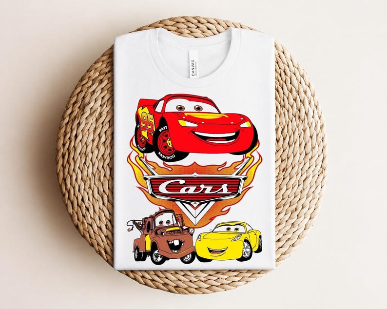 May include: White t-shirt featuring a graphic of the Cars movie characters Lightning McQueen, Mater, and a yellow car. The design includes the word "Cars" in a flame-like font. The shirt is folded on a woven surface.