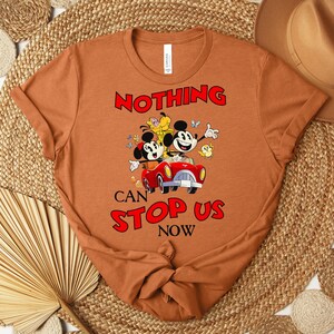 Puede incluir: Camiseta de color &oacute;xido con un gr&aacute;fico de Mickey y Minnie Mouse en un coche rojo, con el texto "NOTHING CAN STOP US NOW" en rojo. El dise&ntilde;o incluye flores y mariposas de dibujos animados. La camiseta se muestra sobre una estera tejida.