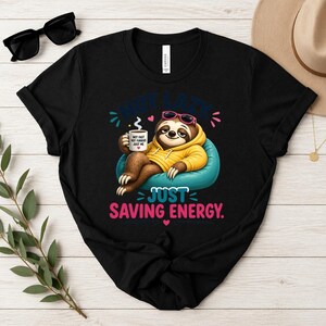 May include: Black t-shirt with a cartoon sloth wearing sunglasses and a yellow hoodie, holding a coffee mug. The text on the shirt reads "NOT LAZY JUST SAVING ENERGY."