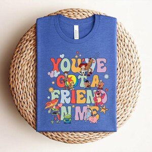 May include: Blue t-shirt with the phrase "You've Got a Friend in Me" in colourful, retro lettering. The shirt features cartoon characters. The t-shirt is made of a soft material.