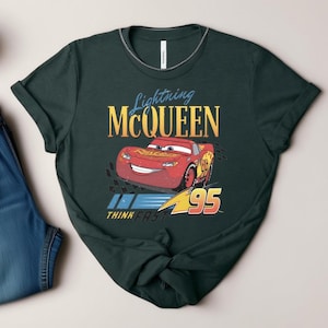 May include: Dark green t-shirt featuring Lightning McQueen from the Cars film. The design includes the text "Lightning McQueen" in gold and blue, the number "95", and the phrase "Think Fast".
