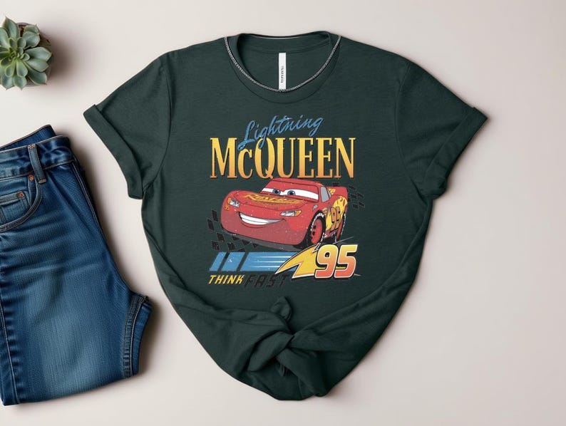 May include: Dark green t-shirt featuring Lightning McQueen from the Cars film. The design includes the text "Lightning McQueen" in gold and blue, the number "95", and the phrase "Think Fast".