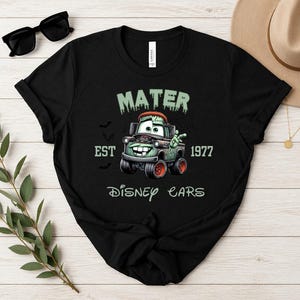 Może przedstawiać: Czarny t-shirt z grafiką Matera z filmu Auta Disneya w stylu zombie. Projekt zawiera napisy "MATER", "EST 1977" i "Disney Cars". Koszulka ma okrągły dekolt i kr&oacute;tkie rękawy.
