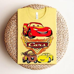 May include: Yellow t-shirt featuring a graphic of Lightning McQueen from the movie Cars, with the word "Cars" in a flame design. The shirt also includes images of Tow Mater and Sally Carrera.