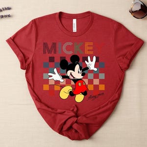 May include: A red t-shirt featuring a graphic of Mickey Mouse with the word "MICKEY" in colorful block letters. Mickey Mouse is depicted with white gloves, a red jacket, and yellow shoes. Sunglasses are visible in the upper right corner.