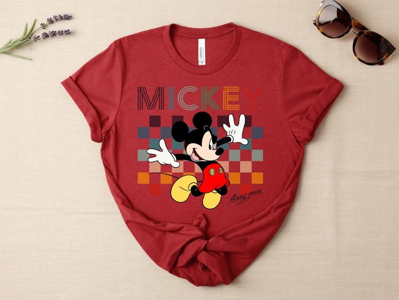 May include: A red t-shirt featuring a graphic of Mickey Mouse with the word "MICKEY" in colorful block letters. Mickey Mouse is depicted with white gloves, a red jacket, and yellow shoes. Sunglasses are visible in the upper right corner.