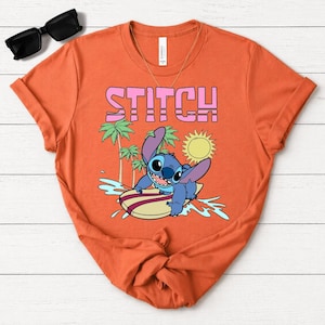 May include: Orange t-shirt featuring the word "STITCH" in pink block letters above a cartoon illustration of Stitch surfing. The design includes palm trees, a sun, and water splashes.