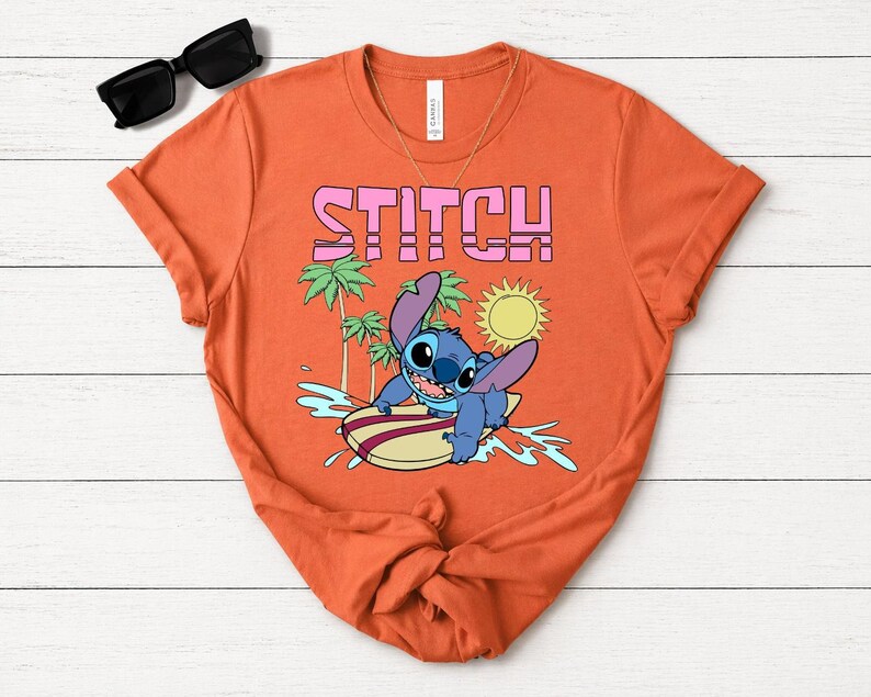 May include: Orange t-shirt featuring the word "STITCH" in pink block letters above a cartoon illustration of Stitch surfing. The design includes palm trees, a sun, and water splashes.