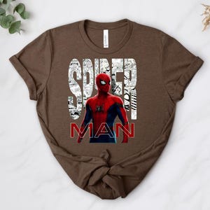 Peut inclure: Un t-shirt marron &agrave; manches courtes avec une illustration du personnage Spider-Man en costume rouge et bleu. Le mot SPIDER est imprim&eacute; en grandes lettres blanches remplies de cases de bande dessin&eacute;e, et MAN est en rouge.