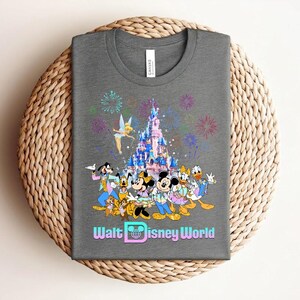 Może przedstawiać: Szara koszulka z kolorową grafiką postaci Disneya, zamkiem, fajerwerkami i napisem "Walt Disney World". Projekt zawiera Myszkę Miki, Minnie, Kaczora Donalda i Goofy'ego.