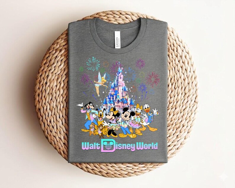Może przedstawiać: Szara koszulka z kolorową grafiką postaci Disneya, zamkiem, fajerwerkami i napisem "Walt Disney World". Projekt zawiera Myszkę Miki, Minnie, Kaczora Donalda i Goofy'ego.
