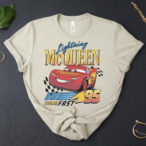 May include: Beige t-shirt with a graphic of Lightning McQueen from the Cars film. The design includes the text "Lightning McQueen", the number "95", and the phrase "Think Fast". The red car is the focal point of the design.