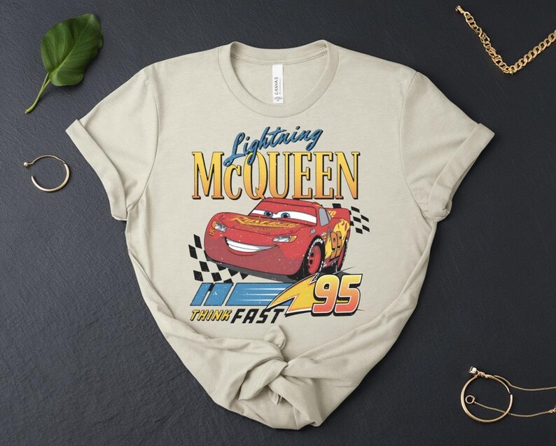 May include: Beige t-shirt with a graphic of Lightning McQueen from the Cars film. The design includes the text "Lightning McQueen", the number "95", and the phrase "Think Fast". The red car is the focal point of the design.