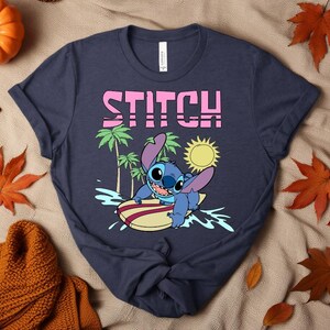 May include: Navy blue t-shirt featuring the word "STITCH" in pink block letters above a cartoon image of Stitch surfing. The image includes palm trees, a yellow sun, and Stitch on a surfboard.