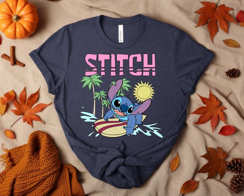 May include: Navy blue t-shirt featuring the word "STITCH" in pink block letters above a cartoon image of Stitch surfing. The image includes palm trees, a yellow sun, and Stitch on a surfboard.