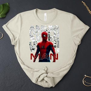 Peut inclure: Un t-shirt beige &agrave; manches courtes avec un graphisme du personnage Spider-Man en costume rouge et bleu. Le mot SPIDER est rempli de panneaux de bande dessin&eacute;e en noir et blanc, et le mot MAN est imprim&eacute; en lettres rouges sous la figure.