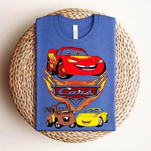 May include: Blue t-shirt featuring a graphic of the animated movie "Cars." The design includes a red race car, a brown truck, and a yellow car, with the word "Cars" in a flame-like font.