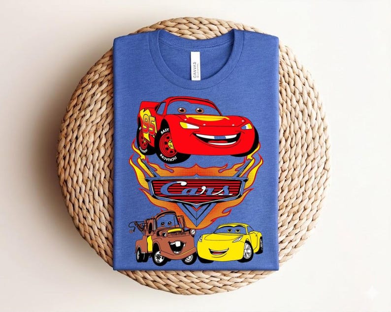 May include: Blue t-shirt featuring a graphic of the animated movie "Cars." The design includes a red race car, a brown truck, and a yellow car, with the word "Cars" in a flame-like font.