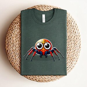 Pu&ograve; includere: T-shirt verde scuro con un disegno di ragno cartoon colorato. Il ragno ha grandi occhi e dettagli rossi, blu e gialli. La maglietta &egrave; piegata su una superficie intrecciata.