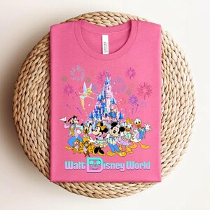 Może przedstawiać: R&oacute;żowa koszulka z kolorową grafiką postaci Disneya, zamkiem, fajerwerkami i napisem "Walt Disney World". Koszulka jest złożona na tkanej, okrągłej powierzchni.