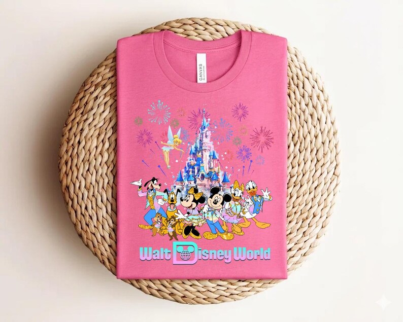 Może przedstawiać: R&oacute;żowa koszulka z kolorową grafiką postaci Disneya, zamkiem, fajerwerkami i napisem "Walt Disney World". Koszulka jest złożona na tkanej, okrągłej powierzchni.