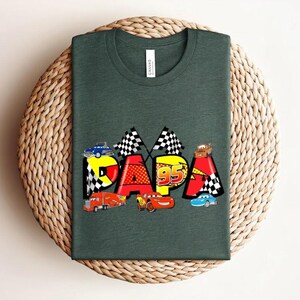 Puede incluir: Camiseta verde oscuro con la palabra "PAPA" en letras coloridas, cada una decorada con coches de carreras y banderas a cuadros. Tambi&eacute;n aparece el n&uacute;mero "95". La camiseta se muestra sobre una superficie circular tejida.