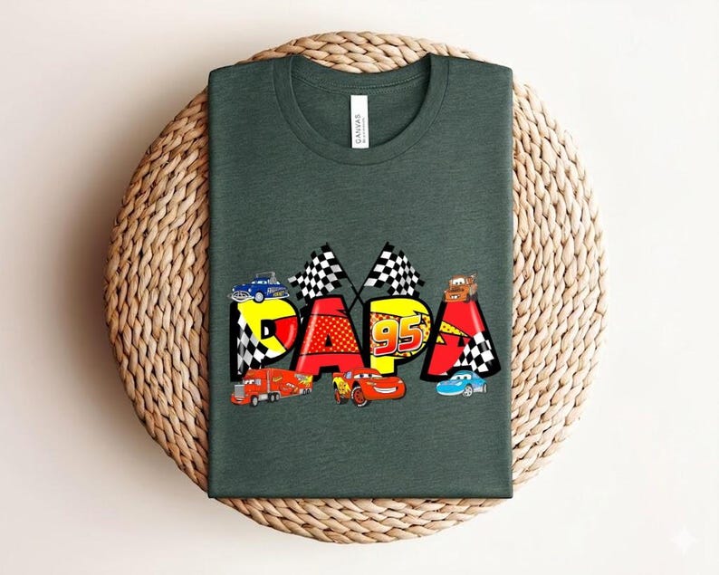 Puede incluir: Camiseta verde oscuro con la palabra "PAPA" en letras coloridas, cada una decorada con coches de carreras y banderas a cuadros. Tambi&eacute;n aparece el n&uacute;mero "95". La camiseta se muestra sobre una superficie circular tejida.