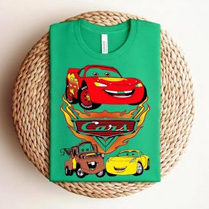 May include: A green t-shirt featuring a graphic of the Cars movie characters. The design includes a red race car, a brown truck, and a yellow car, with the word "Cars" in a stylized font.