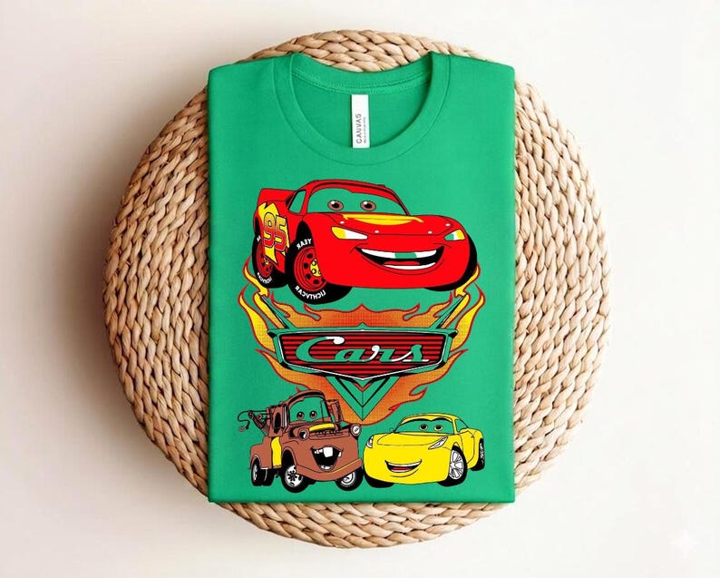May include: A green t-shirt featuring a graphic of the Cars movie characters. The design includes a red race car, a brown truck, and a yellow car, with the word "Cars" in a stylized font.