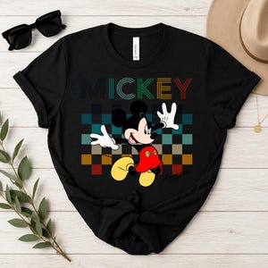 May include: Black t-shirt featuring a colorful graphic of Mickey Mouse with the word "MICKEY" above. Mickey is depicted with his signature red shorts, yellow shoes, and white gloves, set against a checkered background.