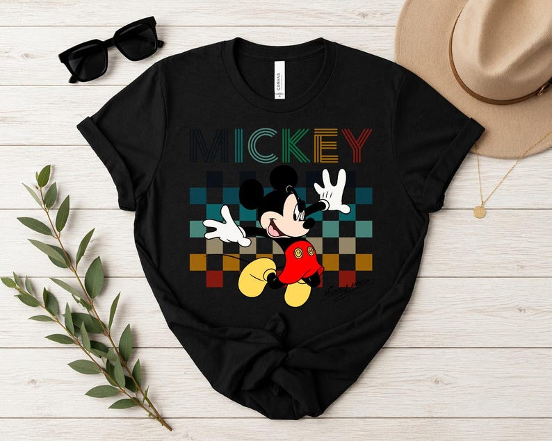 May include: Black t-shirt featuring a colorful graphic of Mickey Mouse with the word "MICKEY" above. Mickey is depicted with his signature red shorts, yellow shoes, and white gloves, set against a checkered background.