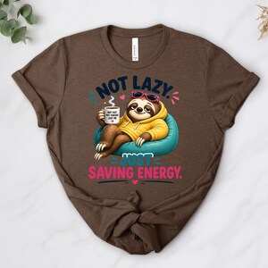 May include: Brown t-shirt with a cartoon sloth wearing sunglasses and a yellow hoodie, relaxing on a blue beanbag chair. The text reads "Not lazy, just saving energy." The sloth is holding a mug.