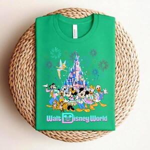 Może przedstawiać: Zielona koszulka z kolorową grafiką postaci Disneya, zamkiem, fajerwerkami i napisem "Walt Disney World". Projekt zawiera Myszkę Miki i Minnie, Kaczora Donalda i Goofy'ego.