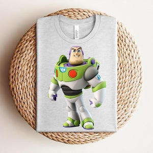 May include: A light gray t-shirt featuring a colorful graphic of Buzz Lightyear from Toy Story. The character is depicted in his signature green, white, and purple spacesuit. The shirt is folded on a woven surface.