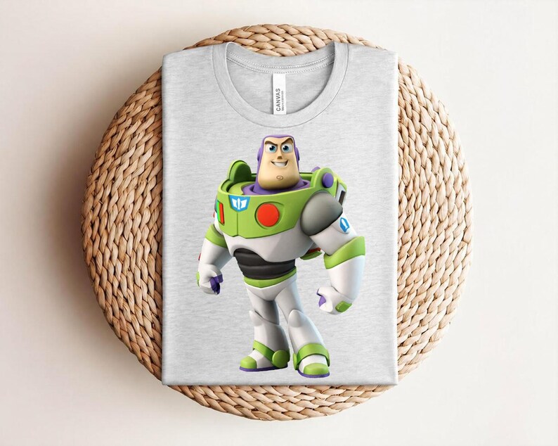 May include: A light gray t-shirt featuring a colorful graphic of Buzz Lightyear from Toy Story. The character is depicted in his signature green, white, and purple spacesuit. The shirt is folded on a woven surface.
