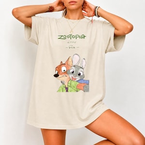 Może przedstawiać: Kremowa koszulka z napisem "Zootopia" w zielonym kolorze i logo Disneya. Poniżej ilustracja Nicka Wilde'a i Judy Hopps. Postacie się obejmują, Nick ma na sobie zieloną koszulę, a Judy niebieski szalik.