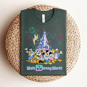 Może przedstawiać: Ciemnozielona koszulka z kolorową grafiką postaci Disneya, w tym Myszki Miki i Minnie, Kaczora Donalda i Goofy'ego, na tle zamku i fajerwerk&oacute;w. Tekst "Walt Disney World" jest wyświetlany poniżej.